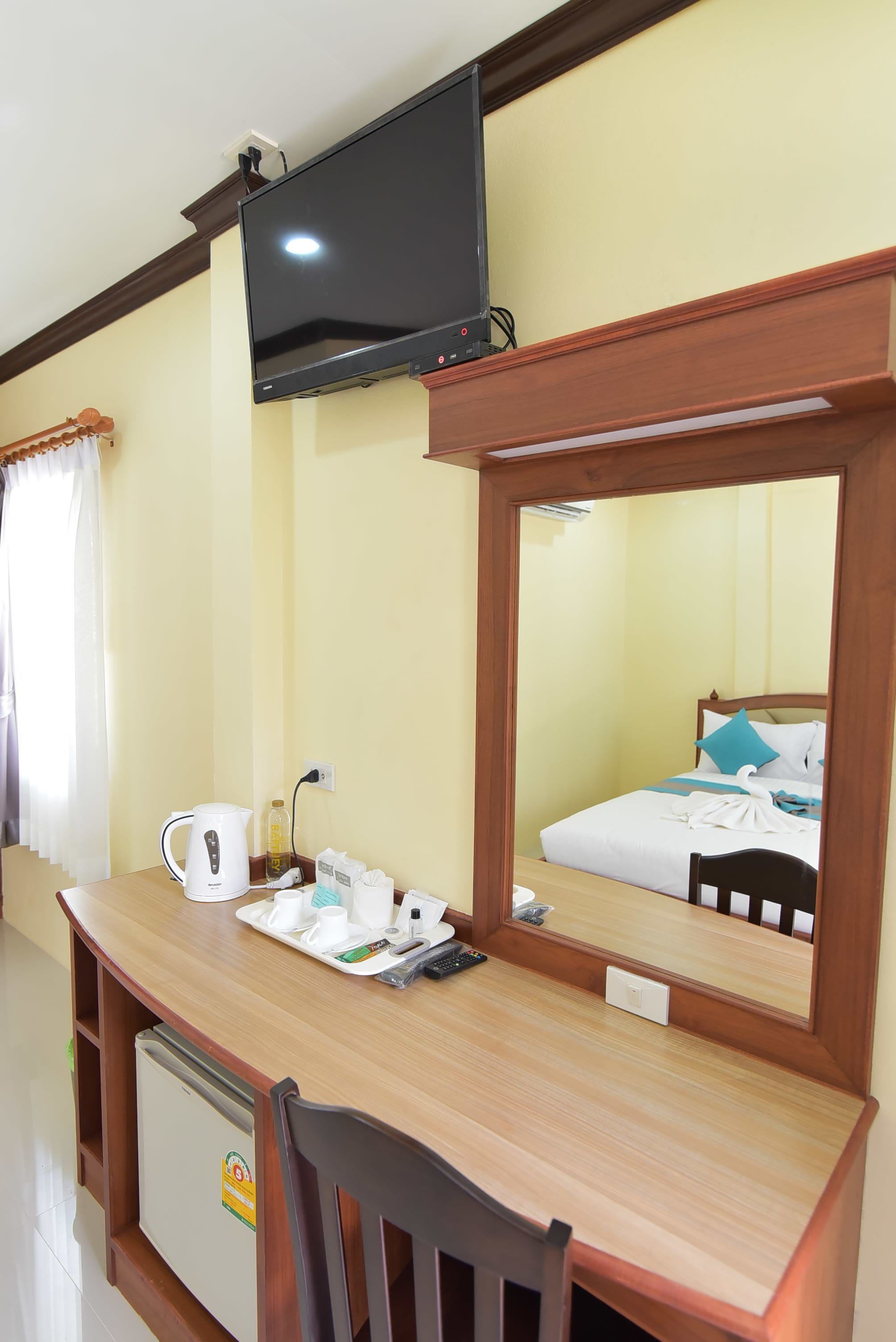 A hotel room with a wooden desk, a television mounted on the wall, a mirror, a bed with blue pillows, and a small refrigerator.