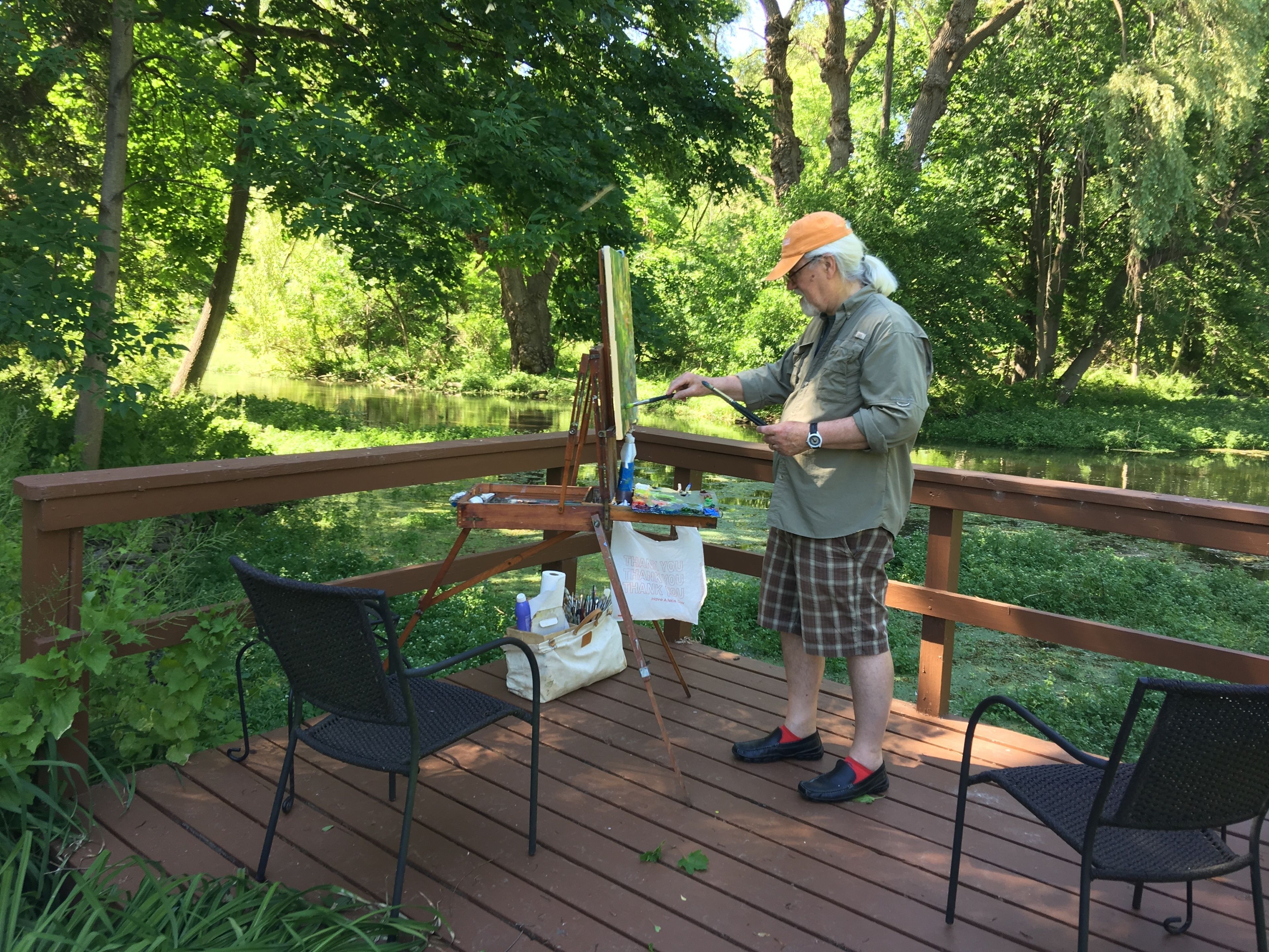 A person painting on an easel on a wooden deck with chairs, surrounded by greenery and a lake.