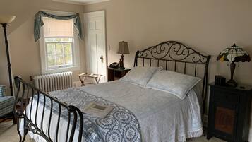 A bedroom with a bed, a nightstand, a lamp, a radiator, a window with curtains, and a door.