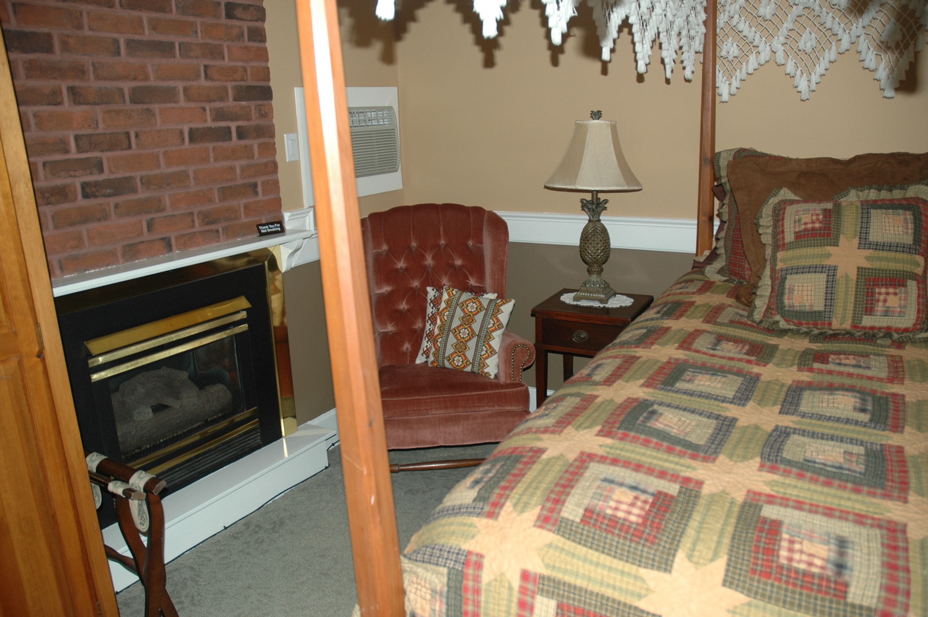 A cozy room with a fireplace, a comfortable armchair, a bedside table with a lamp, and a neatly made bed.