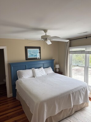 Een slaapkamer met een blauw hoofdeinde, wit beddengoed, een plafondventilator, een ingelijst schilderij en een raam met gordijnen.