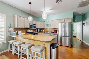 Una cocina con isla central, taburetes de bar, electrodomésticos de acero inoxidable y paredes de color verde azulado.