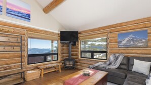 A log cabin interior with a fireplace, wooden furniture, large windows with mountain views, and a cozy seating area.