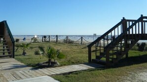 Zona de playa con una escalera de madera que lleva a un acceso a la playa, un pequeño edificio y vistas al océano.