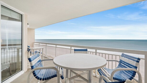 Ein Balkon mit einem weißen Tisch und blauen Stühlen mit Blick auf einen Strand.