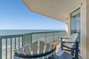A balcony with a view of the ocean, two Adirondack chairs, and a glass door.