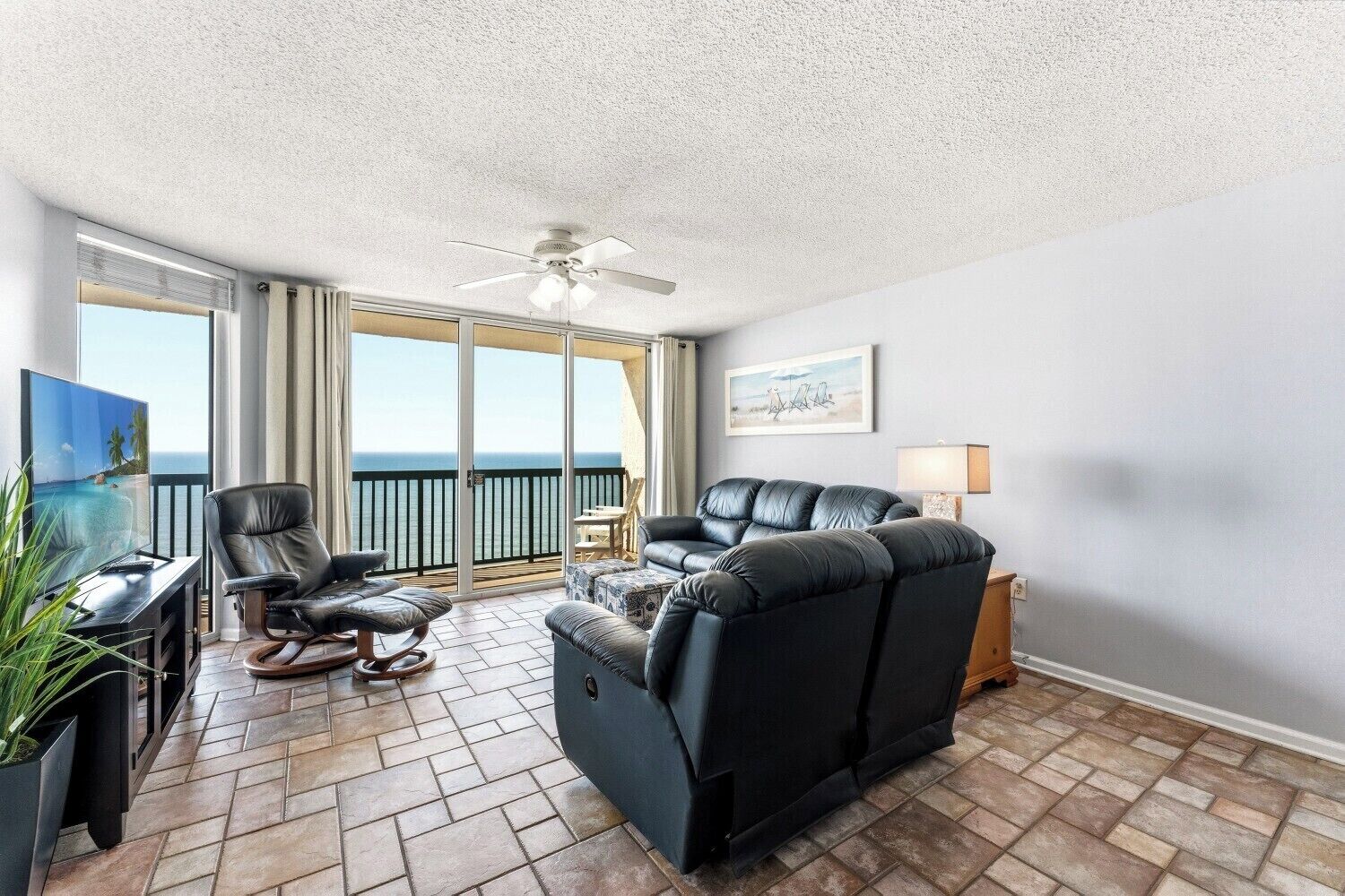 A balcony with a view of the ocean, a ceiling fan, a TV, and a sofa.