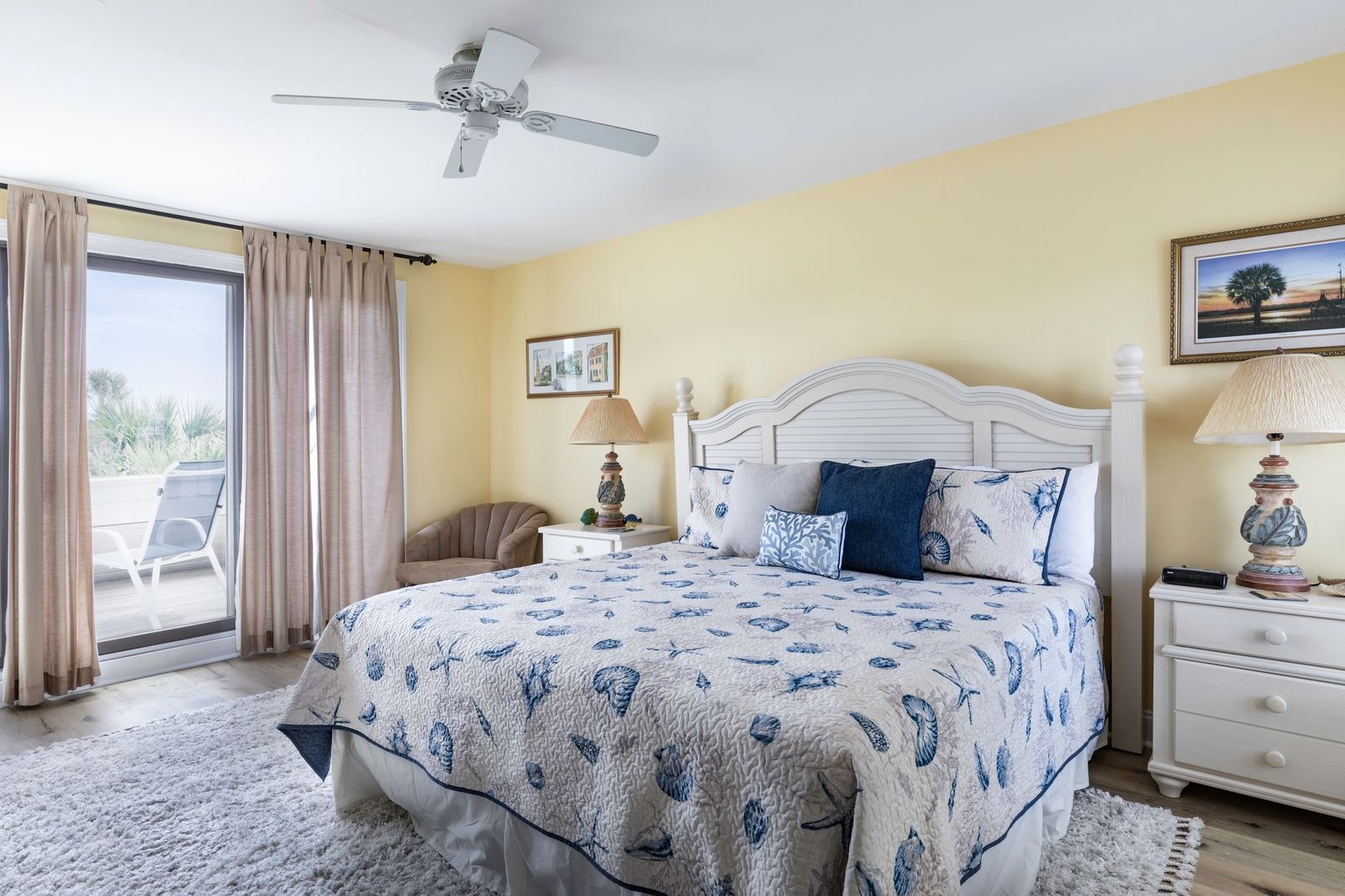 Ein Schlafzimmer mit Bett, Deckenventilator, Nachttisch, Lampe, Stuhl und einem Fenster mit Vorhängen.