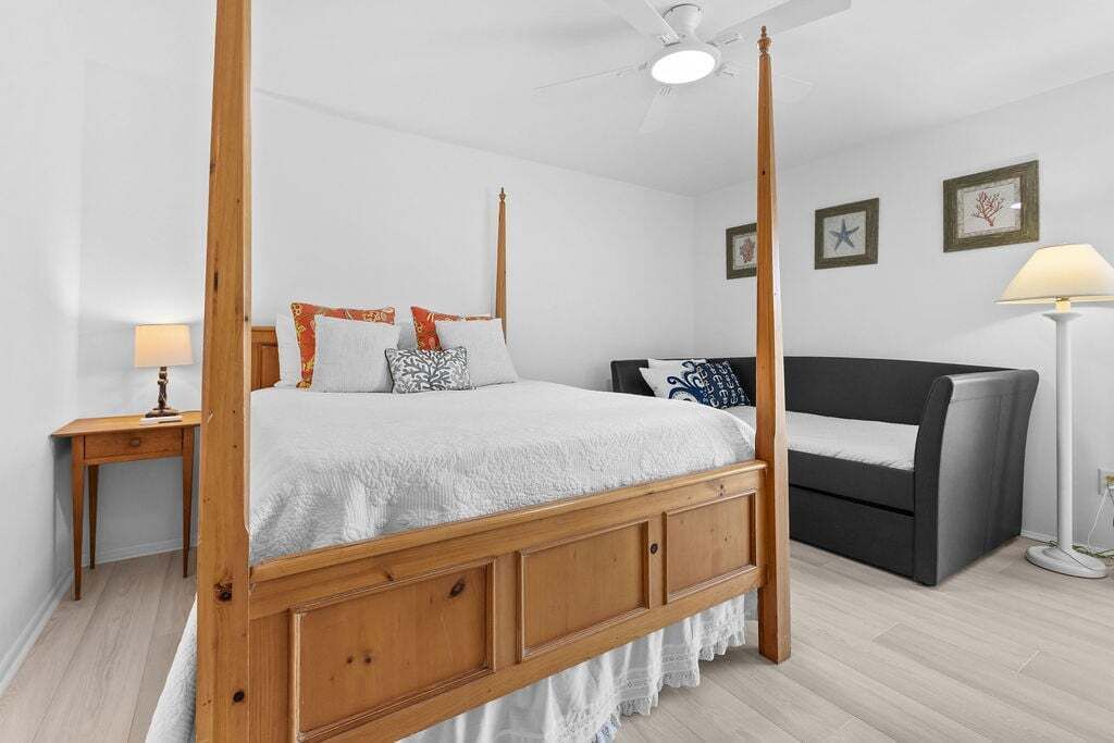 A four-poster bed with a wooden frame, a bedside table with a lamp, a dark sofa, and framed pictures on the wall.