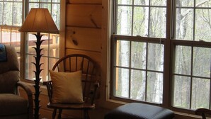 A cabin interior with a high ceiling, large windows, a wooden floor, a table, a chair, and a lamp.