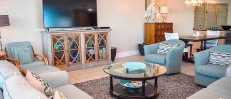 A living room with a flat-screen TV, a glass coffee table, and a dining area with a chandelier.