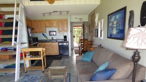 A living room with a sofa, coffee table, and a lamp. There is a kitchen in the background with wooden cabinets and a dining area with a table and chairs. - FULL OCEAN VIEW, 2nd Floor 2 BR/1 BA,  BEST STAND-UP PADDLEBOARDING ON MAUI! (Kihei)