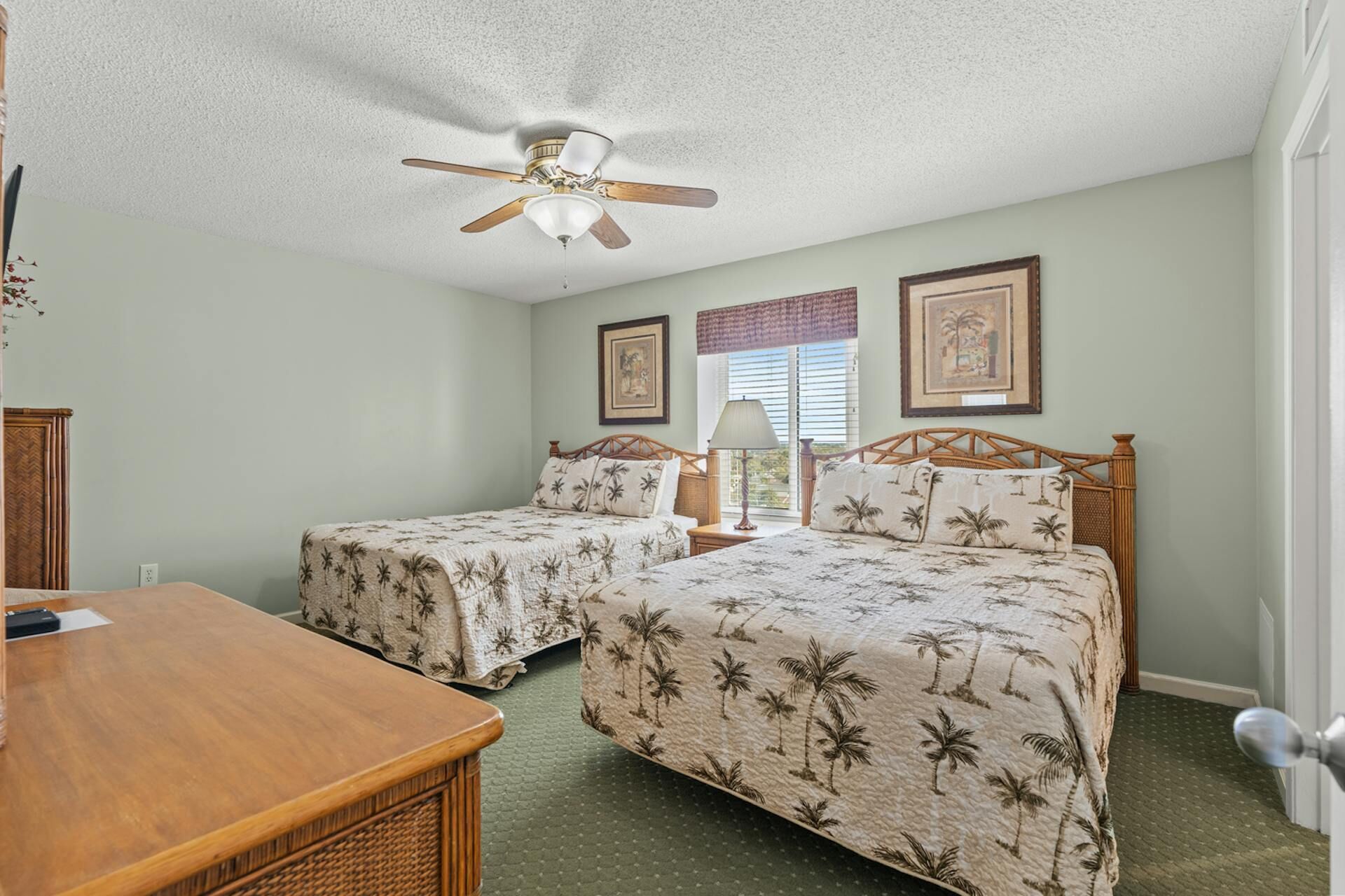 A bedroom with two single beds, a wooden desk, a ceiling fan, and two framed pictures on the wall.
