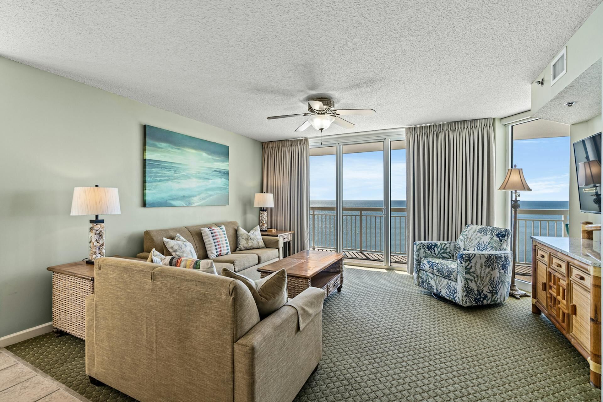 A spacious living room with a large window offering a view of the ocean, a ceiling fan, and a flat-screen TV.
