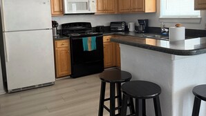 A kitchen with wooden cabinets, a white refrigerator, a black stove, a microwave, a toaster, and a sink. There are three black stools and a window with blinds.