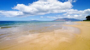 Una playa con agua azul cristalina, una orilla arenosa y montañas lejanas bajo un cielo parcialmente nublado.