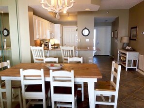 Un coin repas avec une table en bois et des chaises blanches, un lustre et une cuisine en arrière-plan.