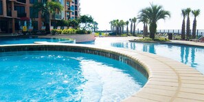 A swimming pool area with a curved edge, surrounded by lounge chairs and palm trees, and a building with balconies in the background. - NEW DISCOUNTS!!! The View! Beautiful, Classy Beach Décor; Very Comfortable! (Orange Beach)