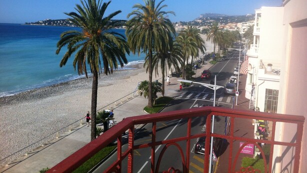 A balcony with a view of a beach, palm trees, and a road with cars and pedestrians. - Blue Riviera: a splendid beachfront 1-bedroom apt w/stunning view (Menton)
