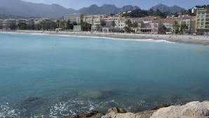 A coastal hotel with multiple floors, a beach in the background, and mountains in the distance. - Blue Riviera: a splendid beachfront 1-bedroom apt w/stunning view (Menton)