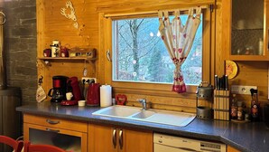 A cozy wooden cabin kitchen with a stone wall, wooden cabinets, a sink, and a dining area with a striped tablecloth. - Chalet Érable**** - Sauna - 8 Pers | Household, Towels & Beds Made Included (Rochesson)