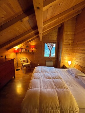 A cozy attic bedroom with a wooden ceiling, a bed, a chair, and a small window with curtains. - Chalet Érable**** - Sauna - 8 Pers | Household, Towels & Beds Made Included (Rochesson)