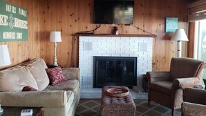 Un salon chaleureux avec une cheminée, des murs en bois, un ventilateur de plafond et un téléviseur fixé au-dessus de la cheminée.