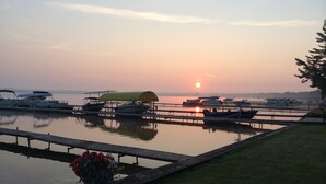 Un port de plaisance avec des bateaux amarrés, une zone herbeuse avec un banc, et un coucher de soleil en arrière-plan.
