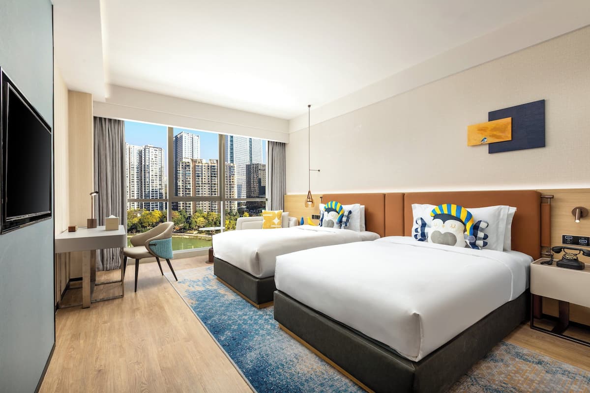 A hotel room with two beds, a desk, and a chair. There is a large window with a view of tall buildings.