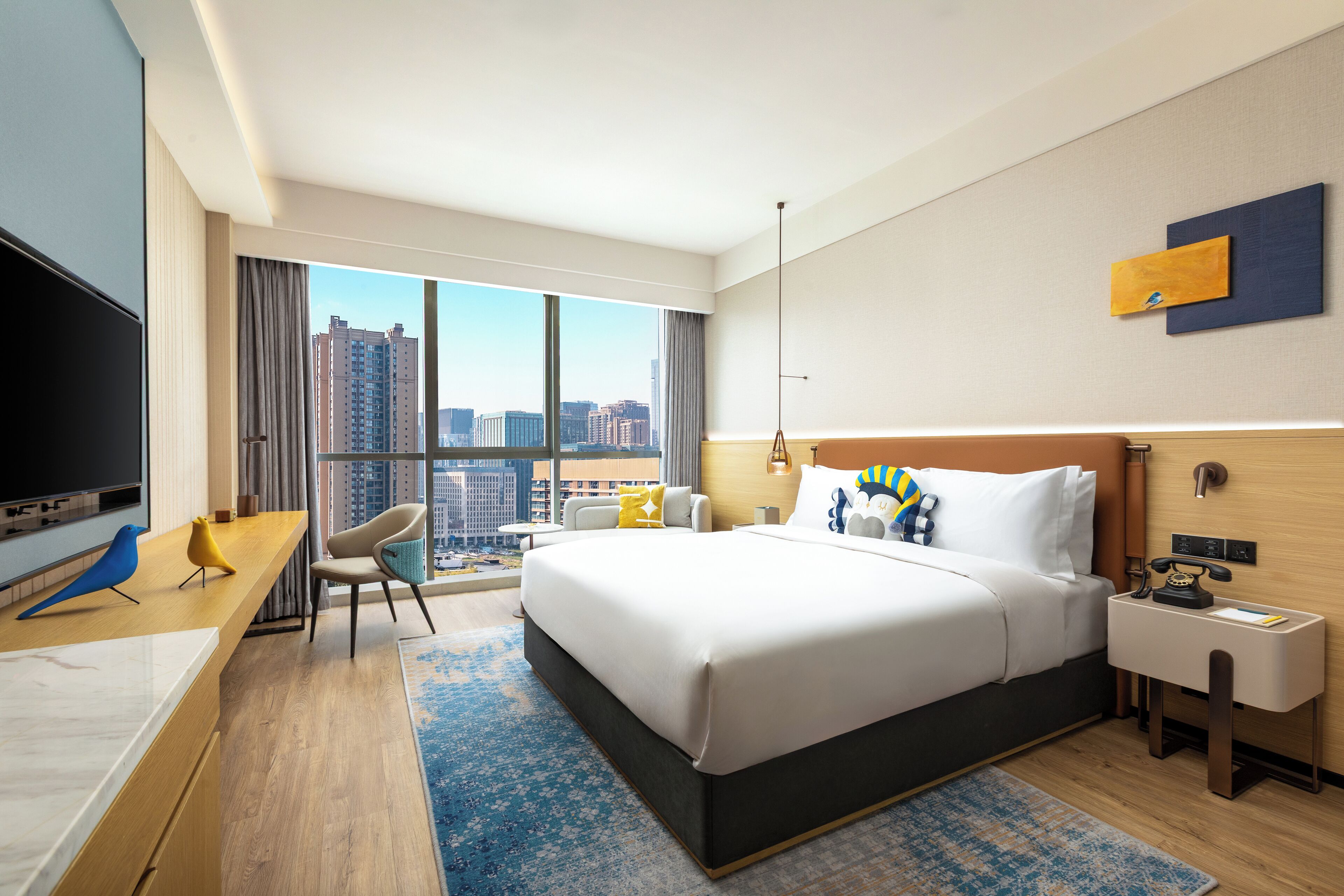A hotel room with a large bed, a desk with a flat-screen TV, a chair, a nightstand, and a view of the cityscape through large windows.
