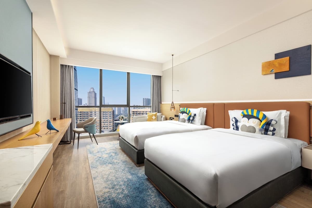 A hotel room with two beds, a TV, a desk, and a view of the city.