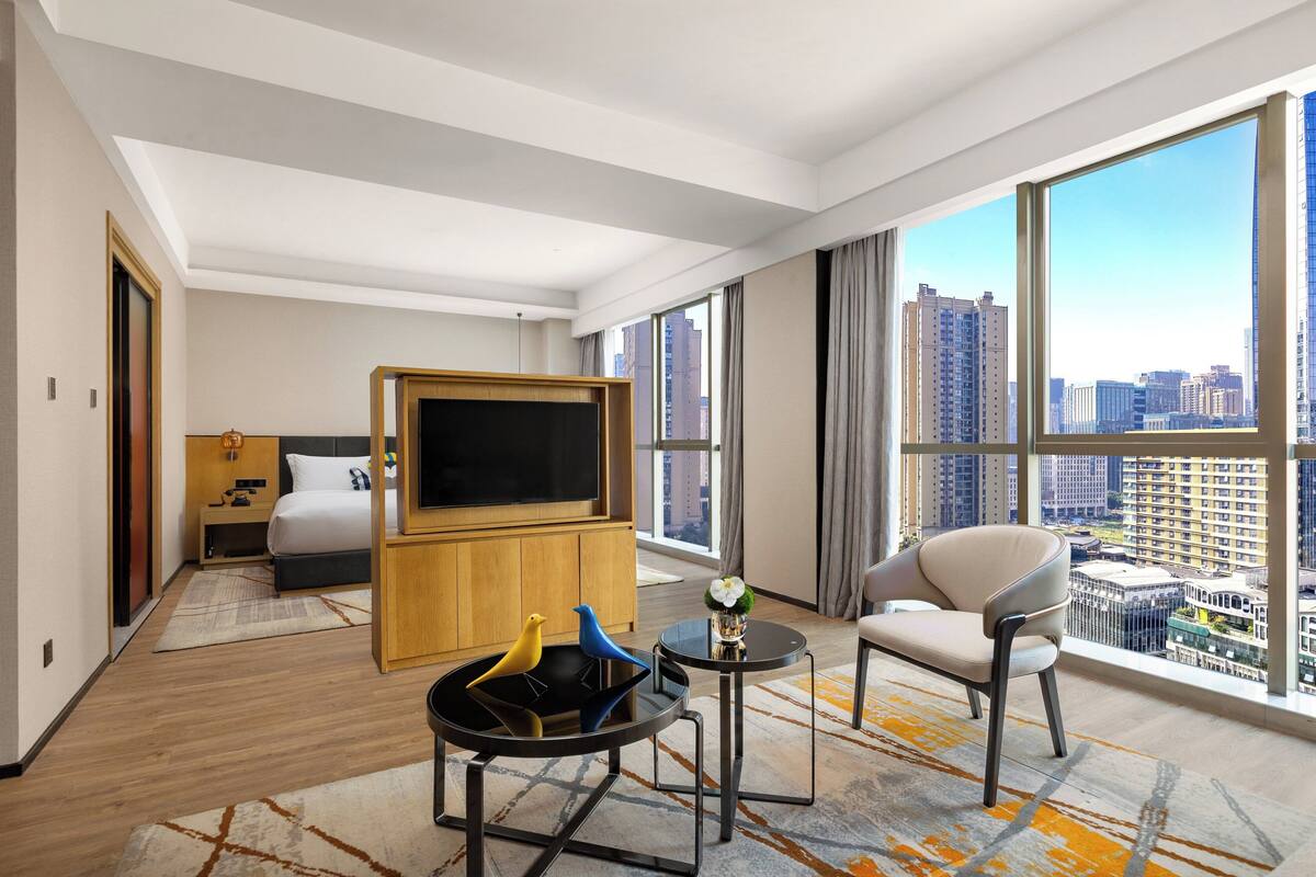 A modern hotel room with a large bed, a TV, a sofa, and a view of the city.