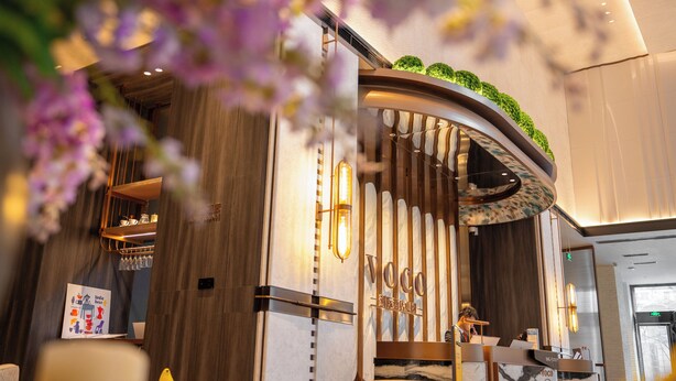 A hotel reception area with a modern design, including a reception desk, a wall-mounted sign, and a decorative plant arrangement.