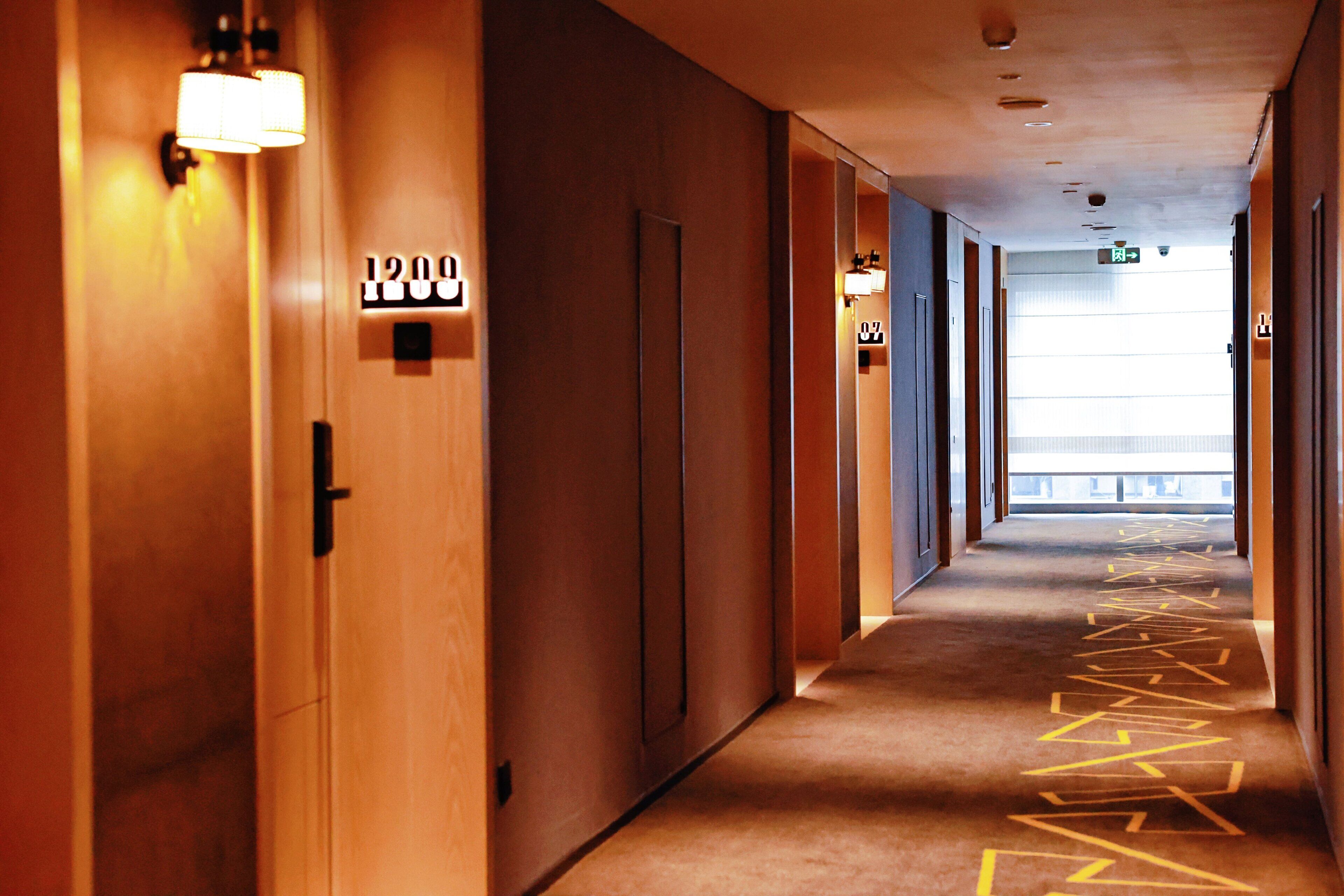 A hotel corridor with wooden doors, numbered room signs, and a carpet with a geometric pattern.