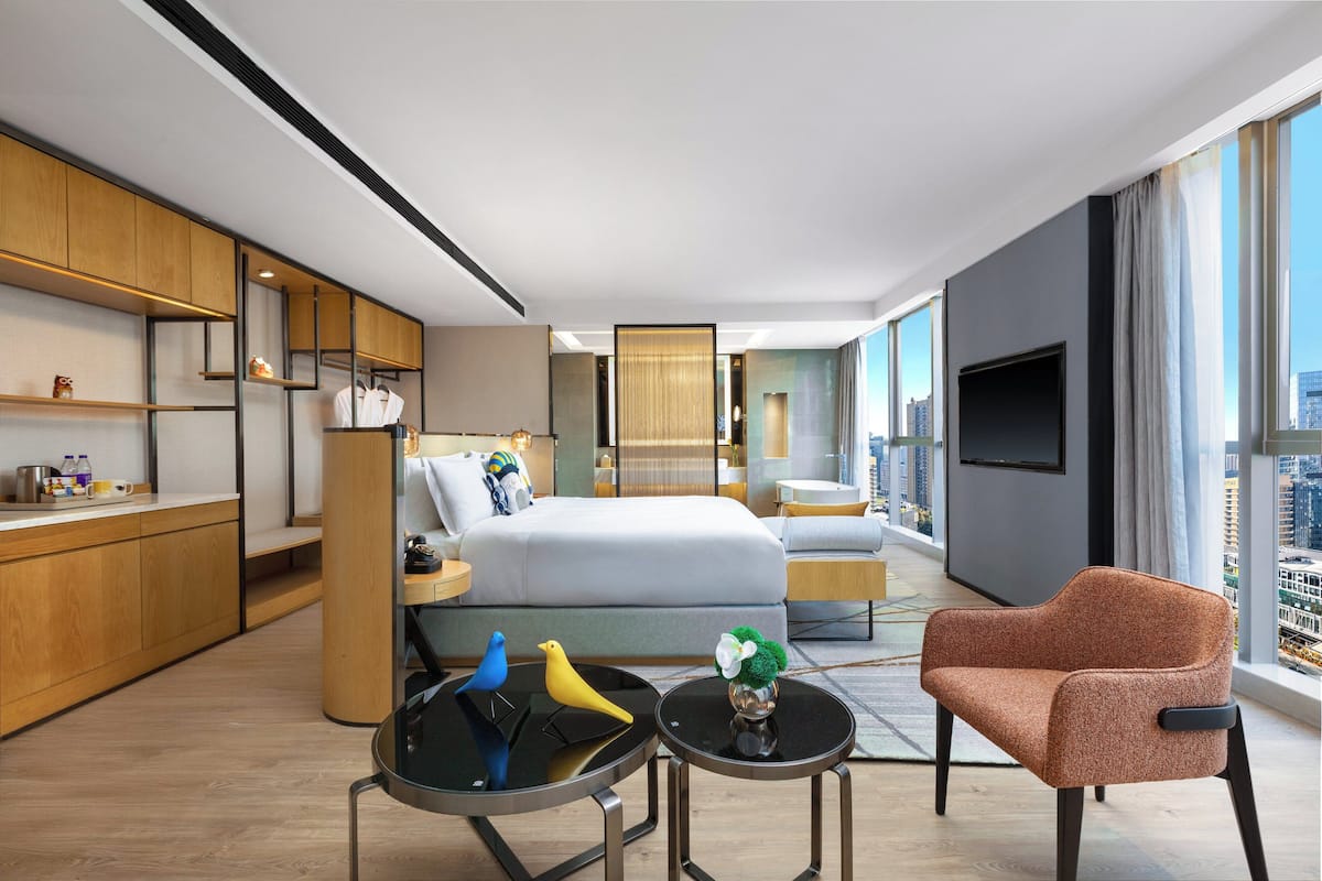 A modern hotel room with a bed, a sitting area with a chair and a small table, a kitchen area with wooden cabinets, and a large window with a city view.