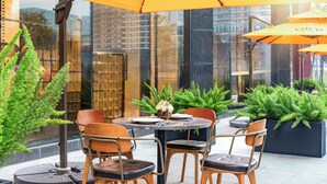 An outdoor seating area with tables and chairs under yellow umbrellas, surrounded by greenery and a city view.