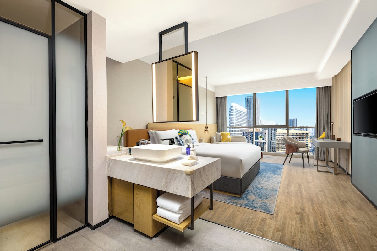 A modern hotel room with a large bed, a desk, a bathroom with a vanity, and a city view.