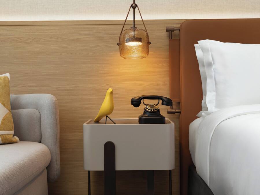 Una habitación de hotel con cama, mesita de noche, lámpara, teléfono y un pájaro.