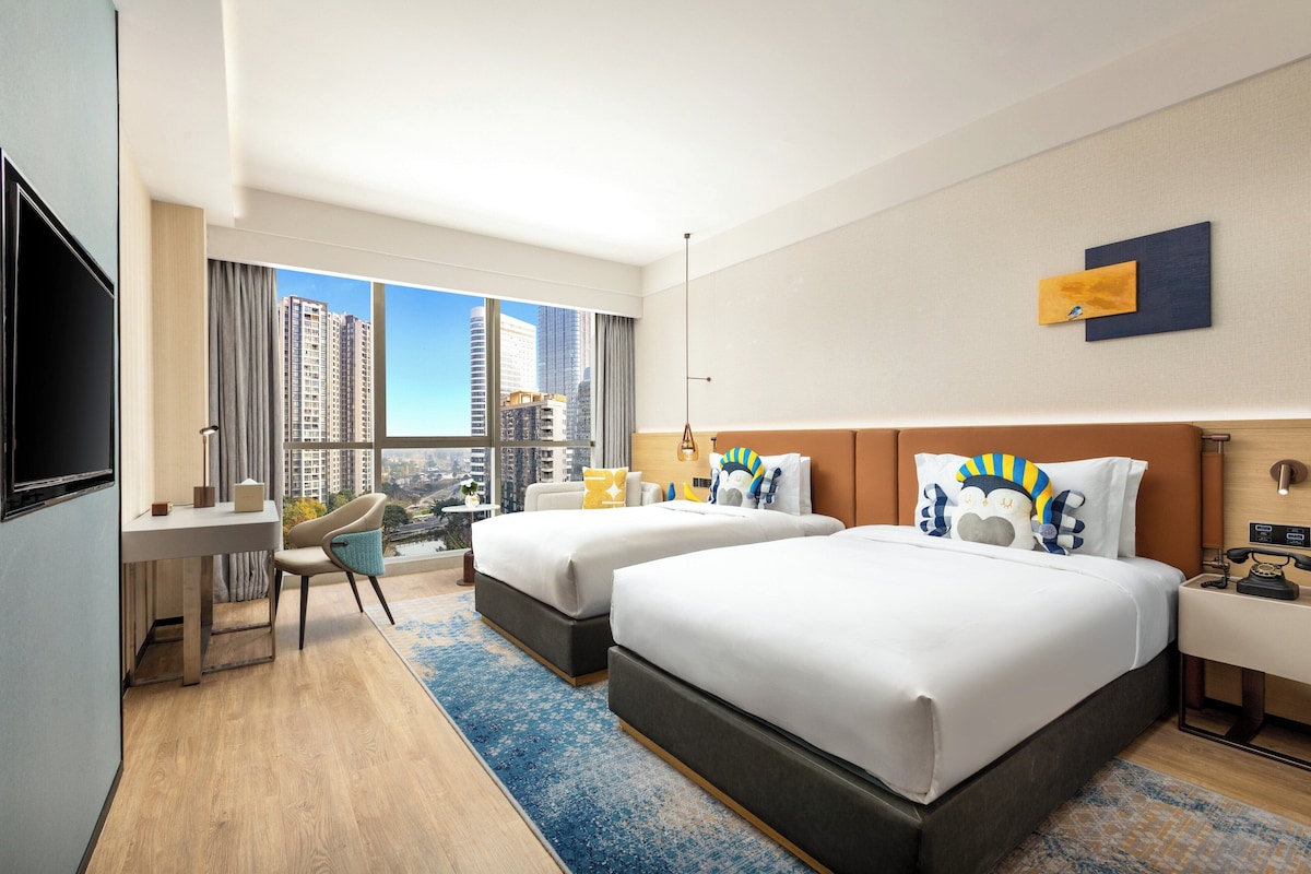 A hotel room with two beds, a desk, a chair, and a large window with a city view.
