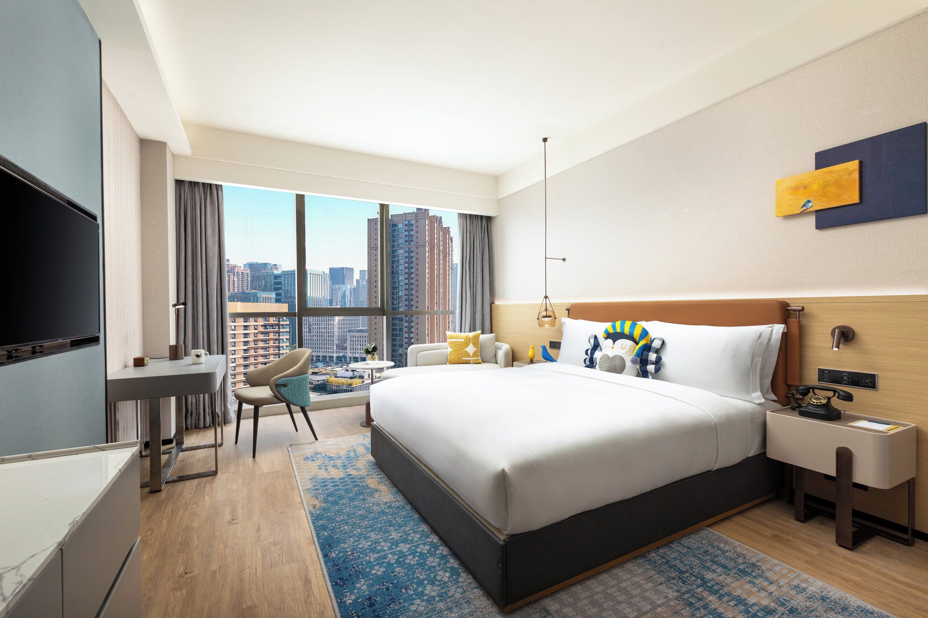 Ein modernes Hotelzimmer mit einem großen Bett, einem Schreibtisch, einem Sessel, einem Fernseher und Blick auf die Stadt.