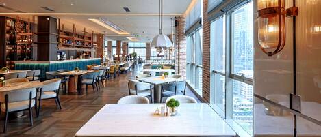 A modern restaurant with a bar area, seating arrangements, and large windows offering a view of the city.