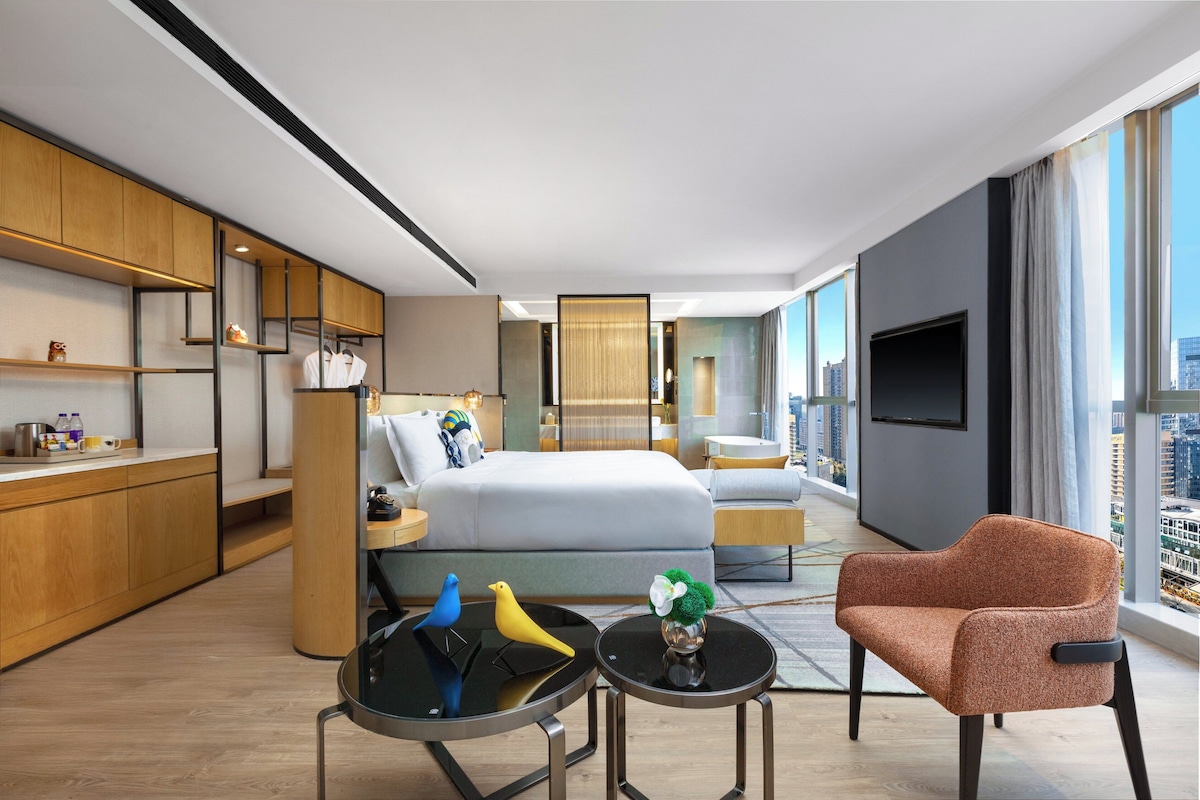 A modern hotel room with a bed, a sitting area with a chair and a small table, a kitchen area with wooden cabinets, and a large window with a city view.