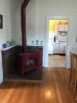 A room with a fireplace, wooden flooring, a kitchen visible through an open door, and a chair. - Water front property with spectacular views. (Marrowstone)