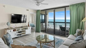 A living room with a TV, sofa, and a glass table. It has a balcony with a view of the ocean. - 9th Floor Oceanfront Condo, Jacuzzi Tub, Outdoor Pool | Ocean Bay Club - 906 (North Myrtle Beach)