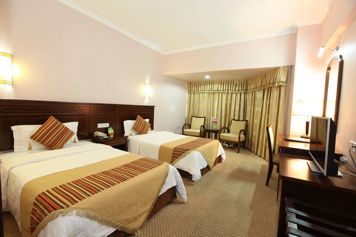 Quarto de hotel com duas camas, uma escrivaninha, uma cadeira, uma televisão e um abajur.