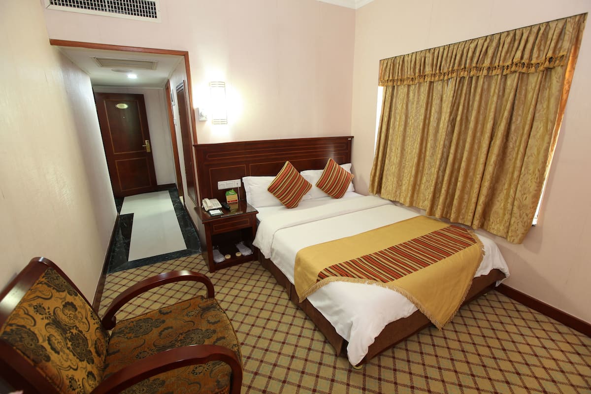Quarto de hotel com uma cama grande, uma mesa de cabeceira de madeira, uma cadeira e vista para o corredor.