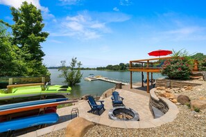 A lakeside patio with kayaks, Adirondack chairs, a fire pit, and a covered seating area with a red umbrella. - Best Lakefront Views, NEW Hot Tub, FirePit, Dock, Kayaks, Dog Friendly (Dandridge)