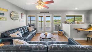 A spacious living room with a black leather sofa, a ceiling fan, and a view of a lake through the glass doors. - Best Lakefront Views, NEW Hot Tub, FirePit, Dock, Kayaks, Dog Friendly (Dandridge)