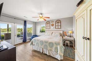 A bedroom with a large bed, a ceiling fan, a view of the outdoors, and a small table with a lamp.
