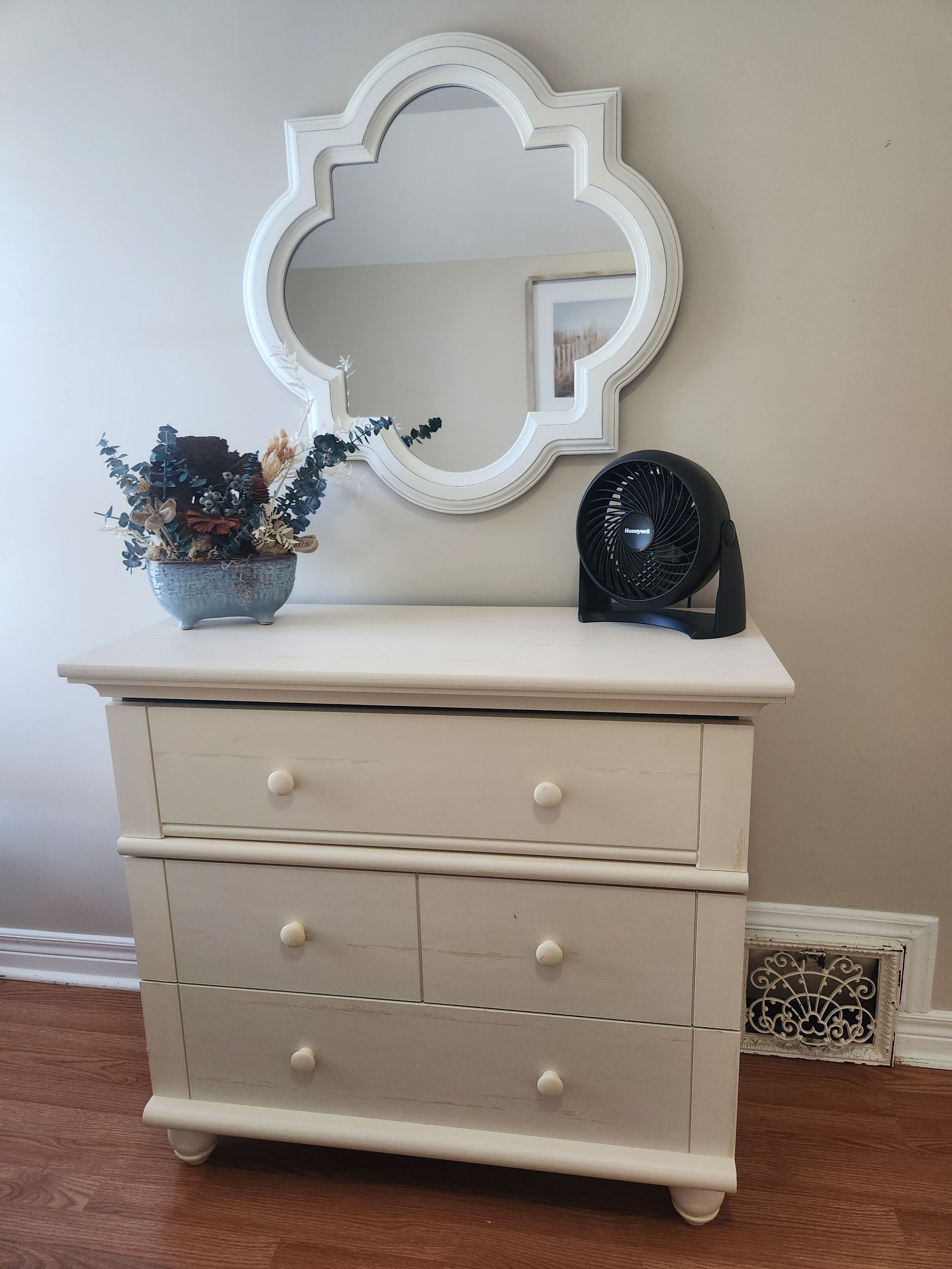 Une commode blanche à trois tiroirs, un miroir décoratif au-dessus, et un petit ventilateur noir posé sur le dessus.